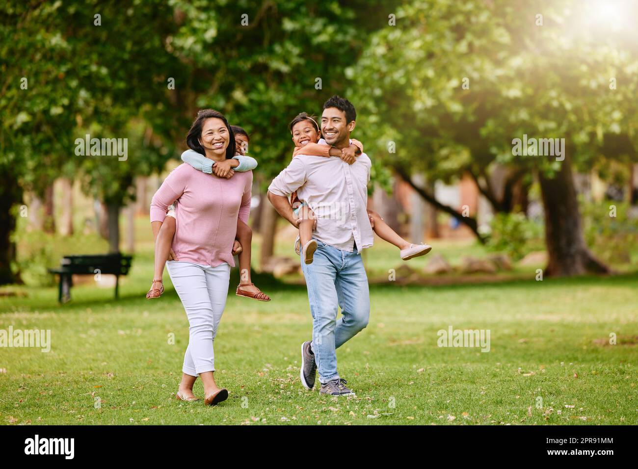 Mixed race family in the park. Happy young mother and father piggybacking their son and daughter outside. Couple carrying their kids while walking on grass during. Two sisters on parents backs Stock Photo