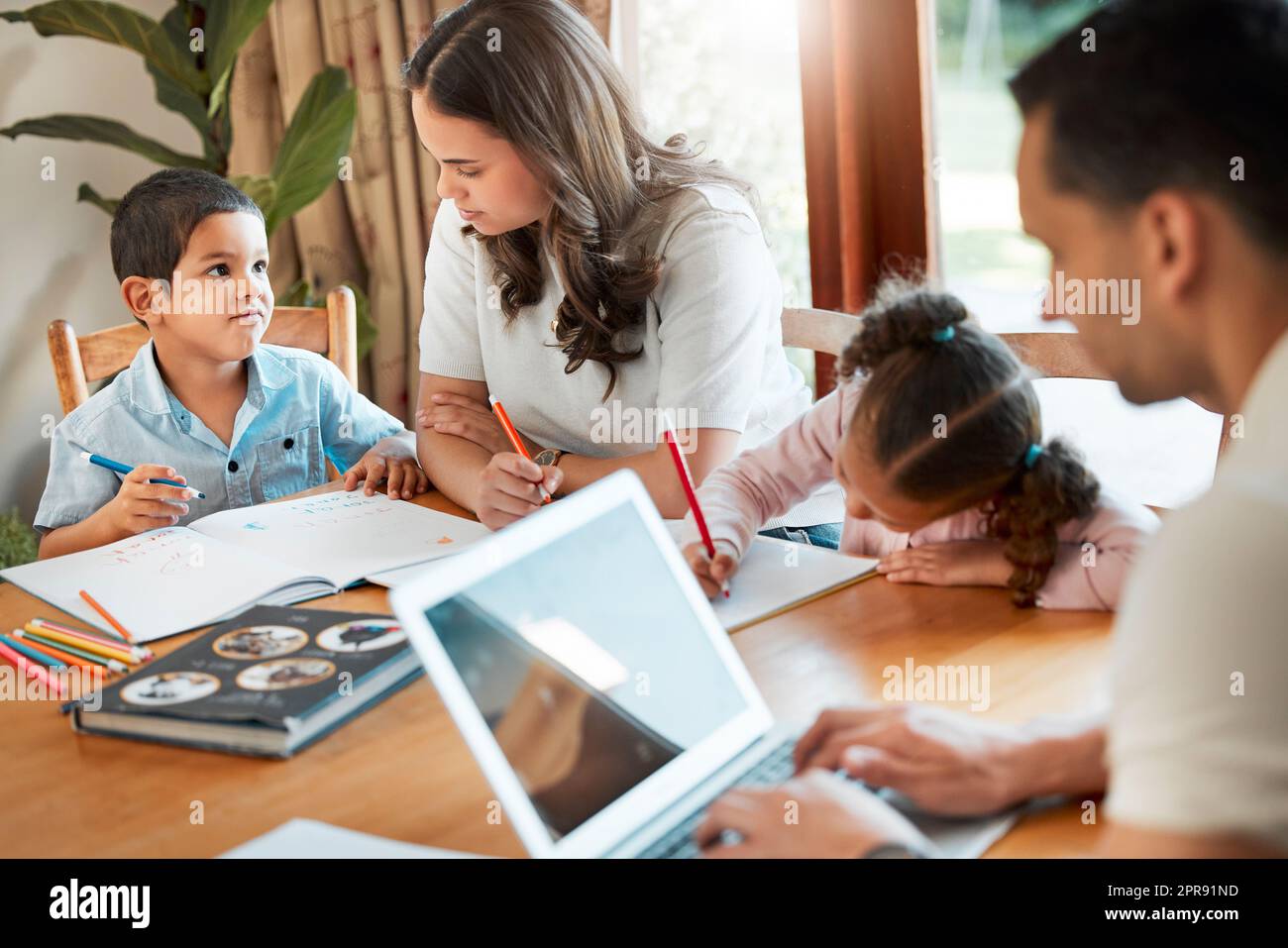 Mother helping her children with their homework. Businessman working from home on his laptop with family. Children being homeschooled in quarantine by parents. Brother and sister drawing pictures Stock Photo