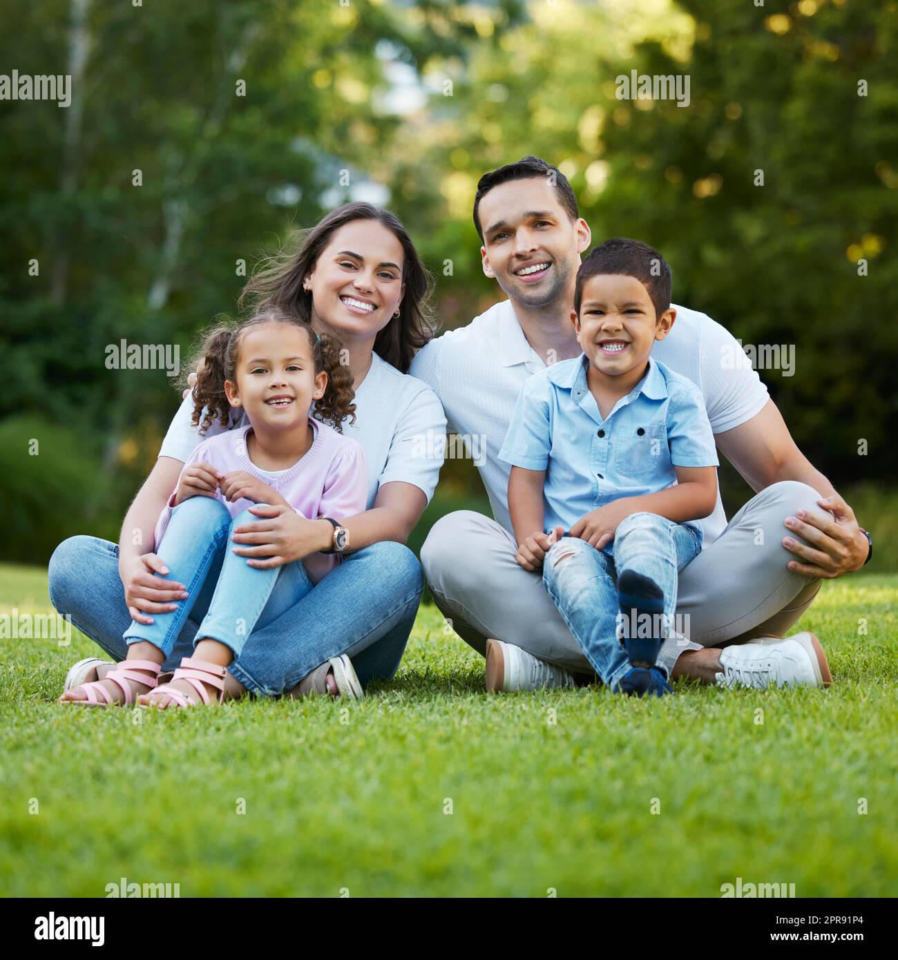 Young happy mixed race family relaxing and sitting on grass in a park together. Loving parents spending time with their little children in a garden. Carefree siblings bonding with their mom and dad Stock Photo