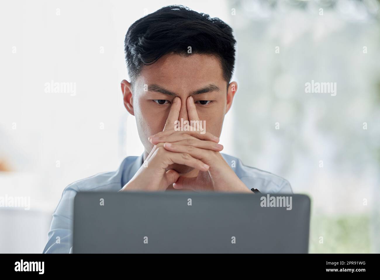 Stressed asian businessman using an office laptop. Thinking professional sitting alone and browsing the internet on technology. Anxious man searching for solution while suffering from a bad headache Stock Photo