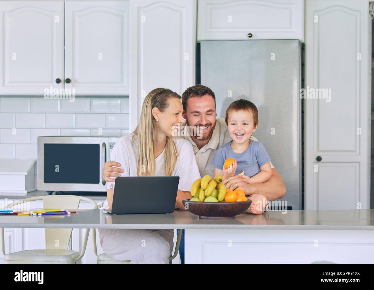 Happy family relaxing in the kitchen. Little boy eating orange in the kitchen. Cheerful caucasian family at home. Father hugging wife and son. Two parents bonding with their son. Young family laughin Stock Photo