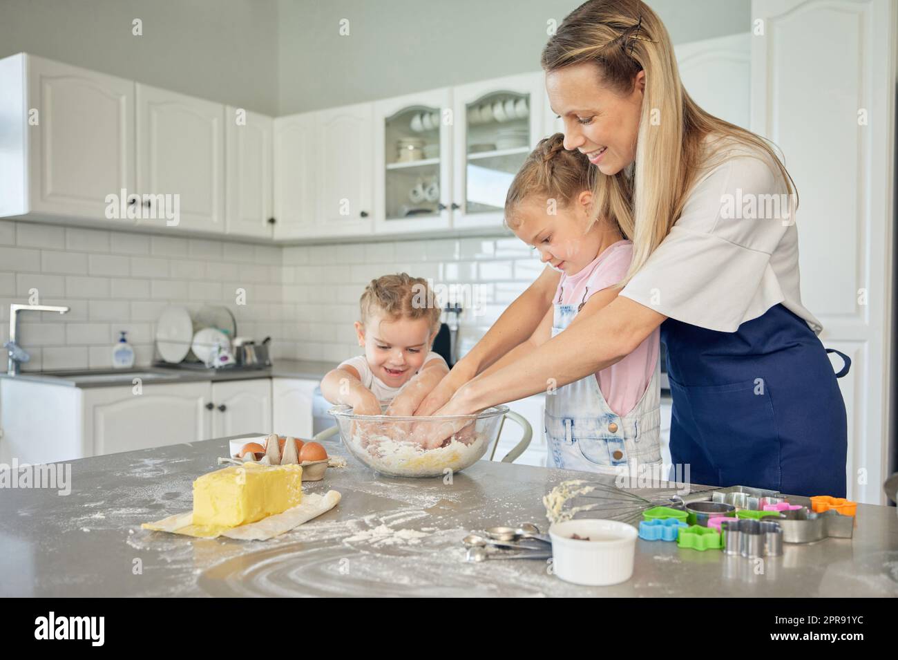 Caucasian caring mother and little daughters baking together in a kitchen at home. Mom teaching girls how to make dough in a messy kitchen. Sisters learning how to bake with their mom Stock Photo