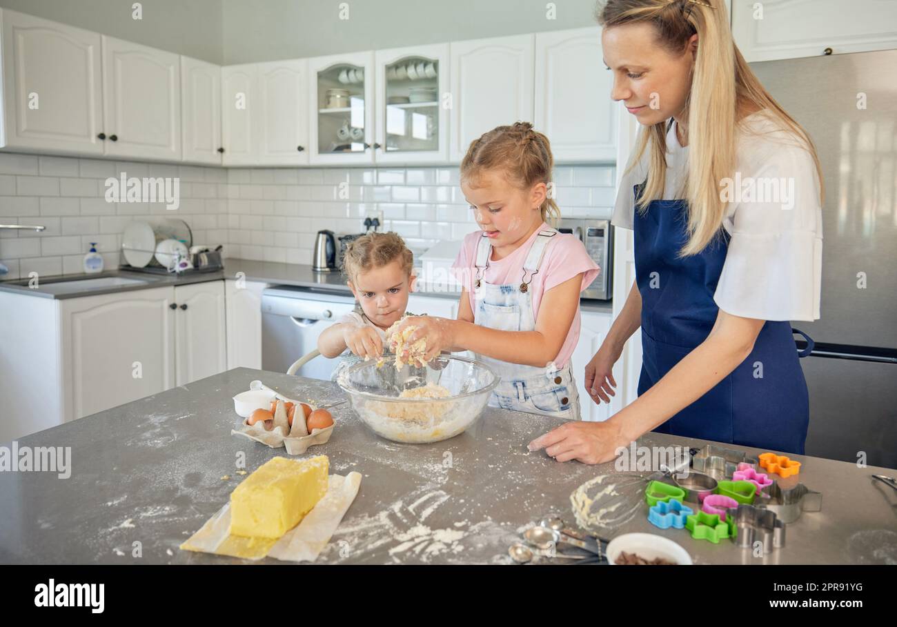 Caucasian mother and little daughters baking together in a kitchen at home. Mom teaching girls how to make dough in a messy kitchen. Sisters learning how to bake with their mom Stock Photo