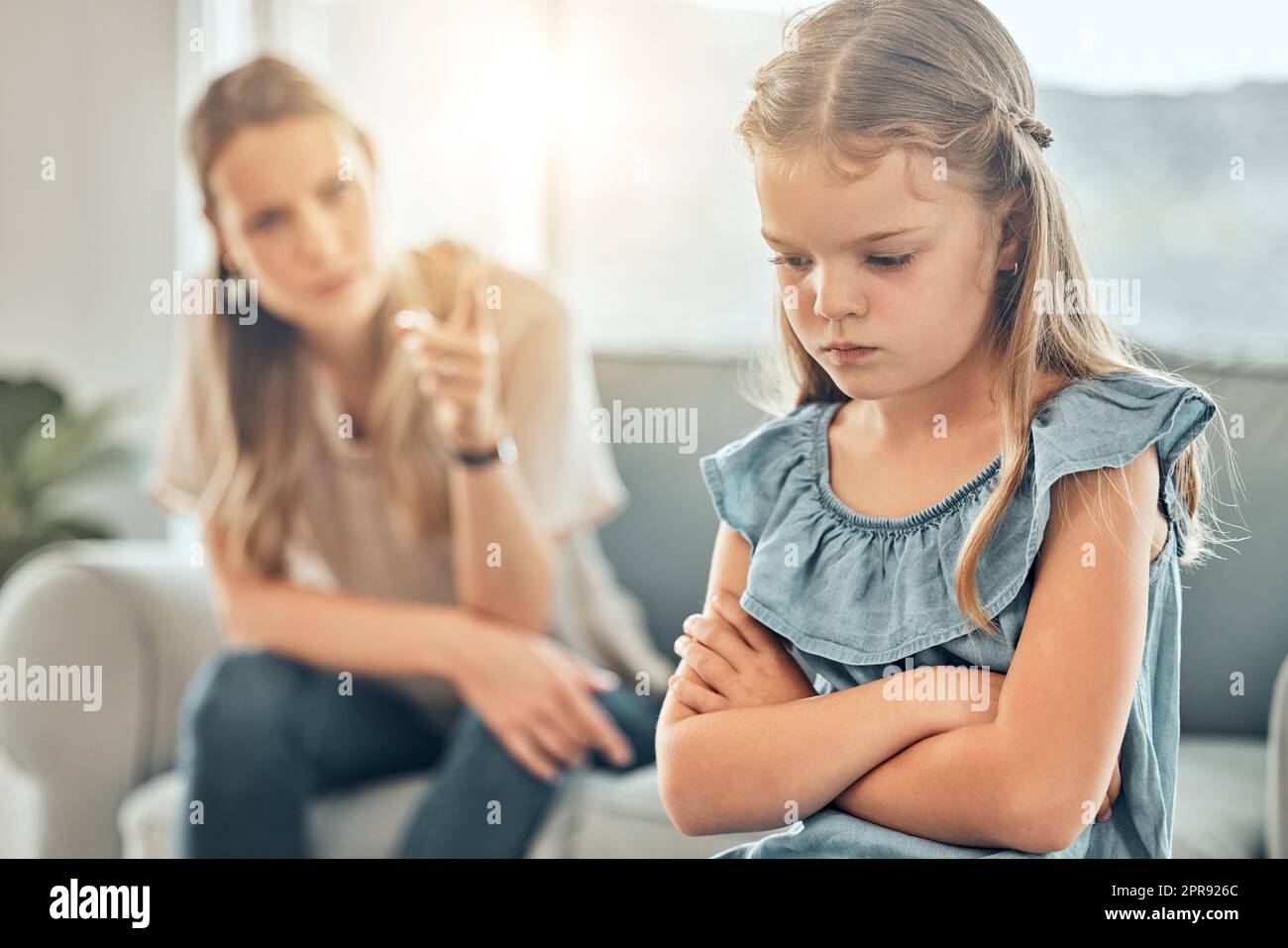Closeup of an adorable little girl standing with arms crossed and looking upset while being scolded and reprimanded by her angry and disappointed mother at home. A woman punishing her young daughter Stock Photo