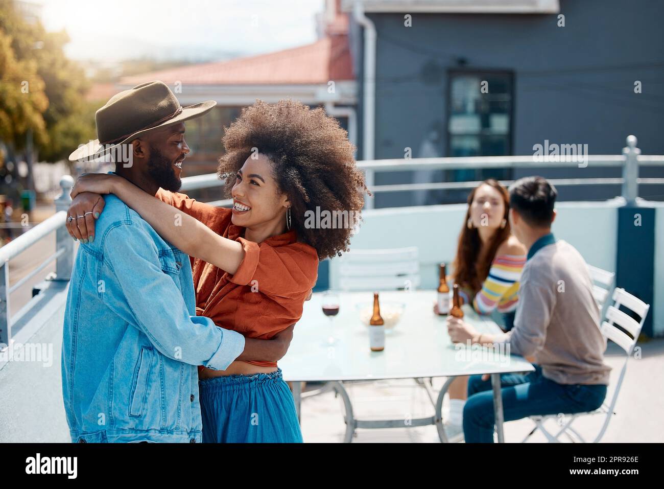 Happy interracial couple hugging while spending time together outside at a restaurant with friends. African american man and mixed race woman embracing while having fun on the weekend Stock Photo