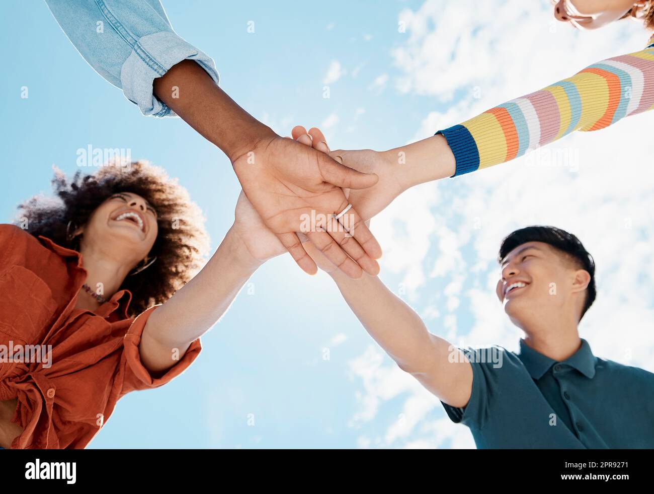 A low close up view of a group of diverse young friends joining hands in a huddle while smiling with a blue sky in the background on a sunny day. Mixed race female with a cool afro hairstyle and her Stock Photo
