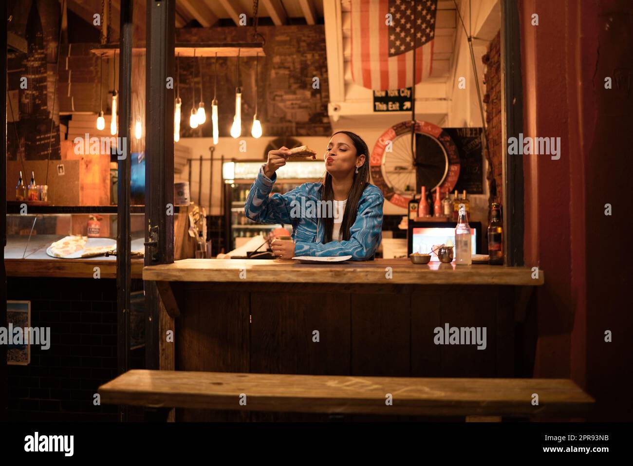 A young and happy woman eating pizza sitting alone in an empty and dark American restaurant. A female relaxing and enjoying delicious food late at night in a cosy junk food, takeaway cafe Stock Photo