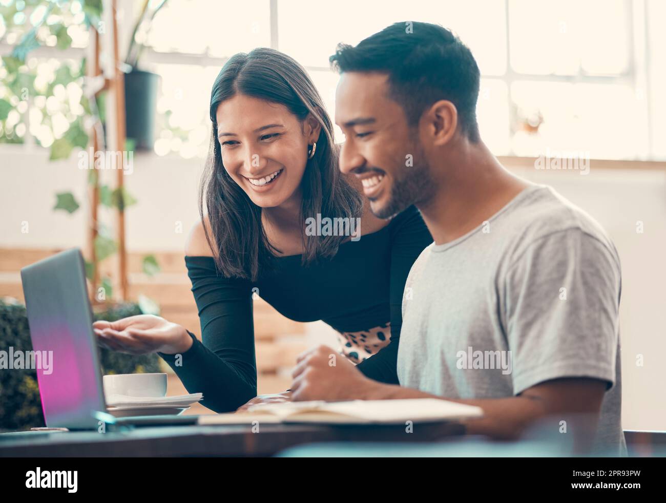 Happy, smiling, cheerful entrepreneurs browsing the internet on a laptop inside a coffee shop. Two latino bloggers sharing ideas and laughing while posting online content inside a quiet restaurant Stock Photo