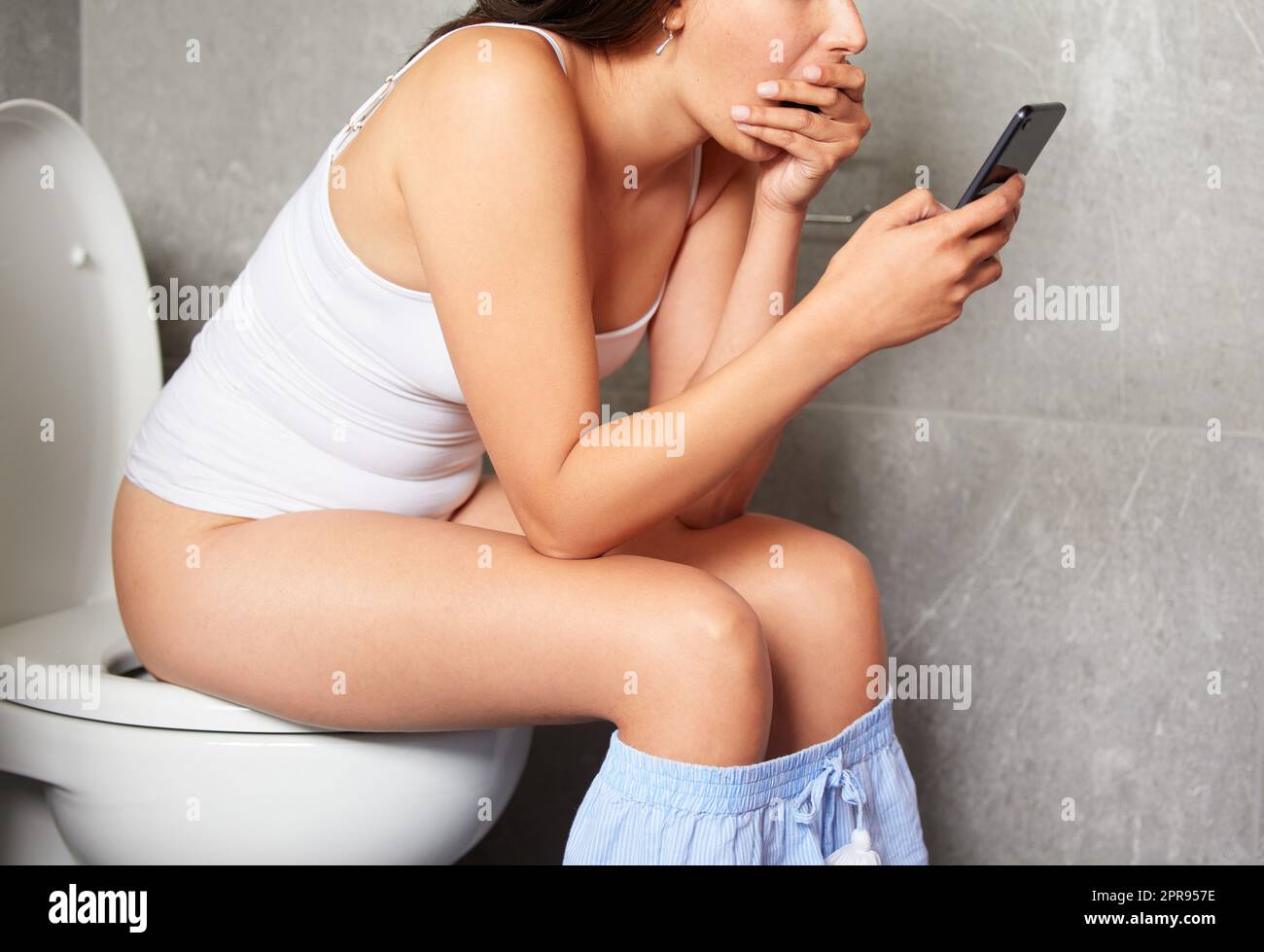 Thats shocking. a young woman using a phone while sitting on the toilet in a bathroom. Stock Photo