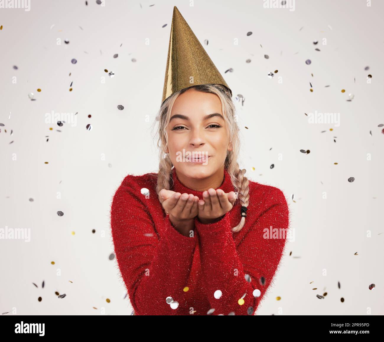 The world has grown weary through the years, but at Christmas it is young. Studio shot of a young woman wearing a party hat and blowing confetti against a grey background. Stock Photo