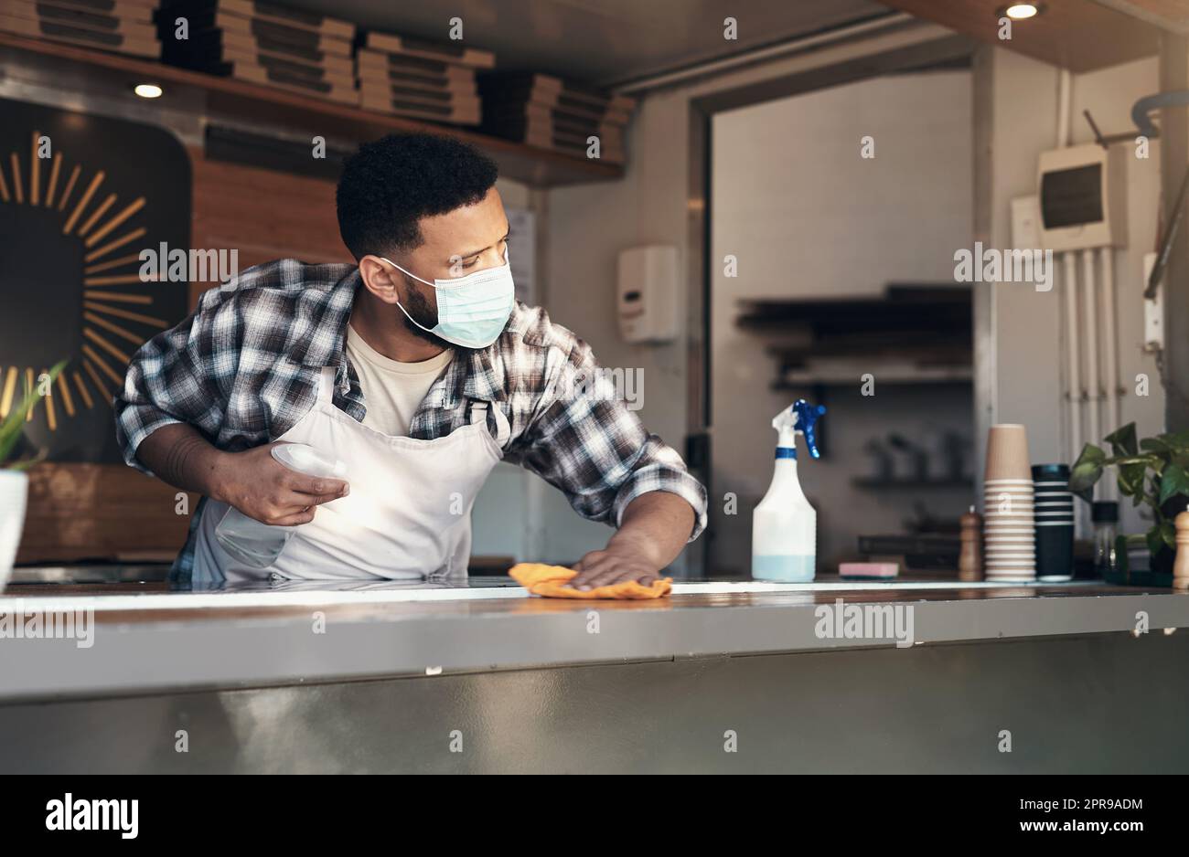 Covid germs are not allowed in my cafe. a young man standing alone and sanitising the counter tops in his restaurant while wearing a face mask. Stock Photo