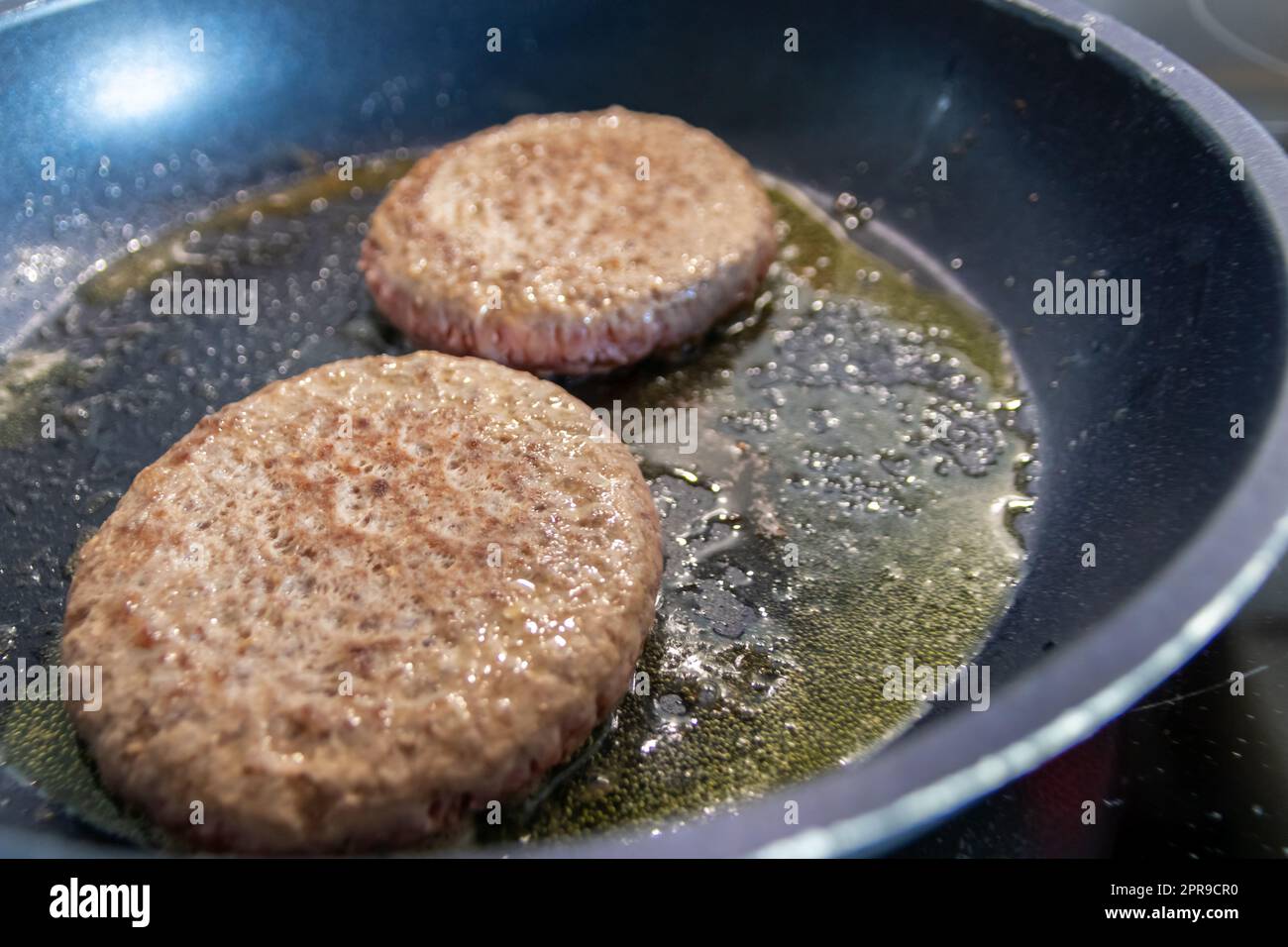 Two burger patties burger meat sizzling in hot pan with fat and oil as delicious selfmade hamburger bbq meatballs as unhealthy fast food lunch with lots of calories and cholesterol in frying pan Stock Photo