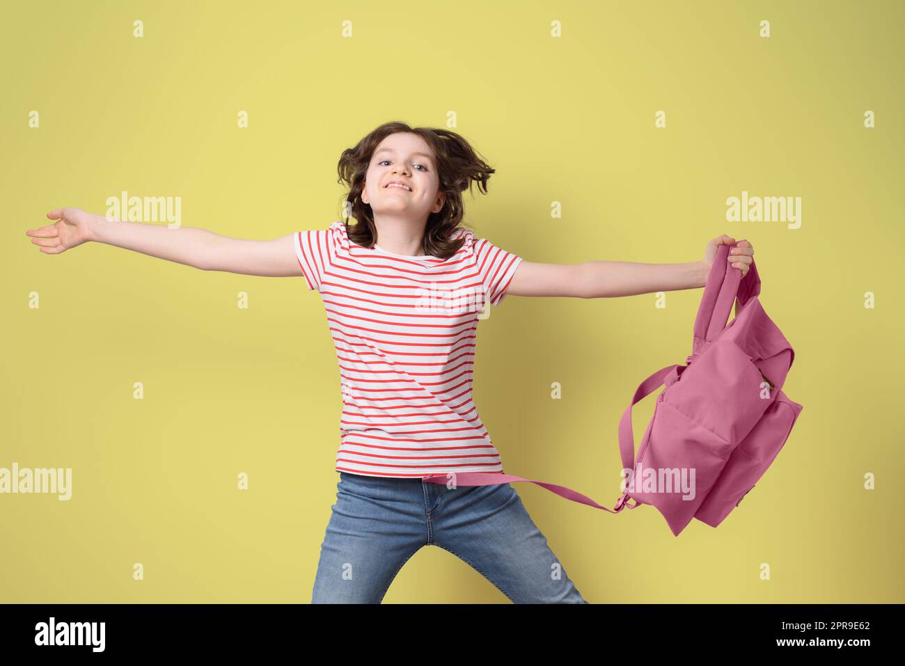 Happy schoolgirl smiling and jumping with pink backpack, looking at camera, on yellow background Stock Photo