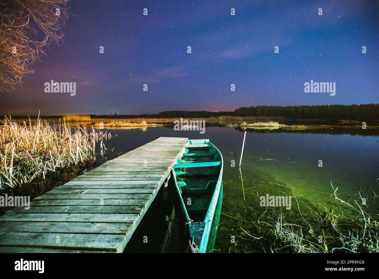 Real Night Sky Stars Above Old Pier With Moored Wooden Fishing Boat. Natural Starry Sky And Countryside Landscape With Lake River In Early Spring Night. Russian Nature Stock Photo