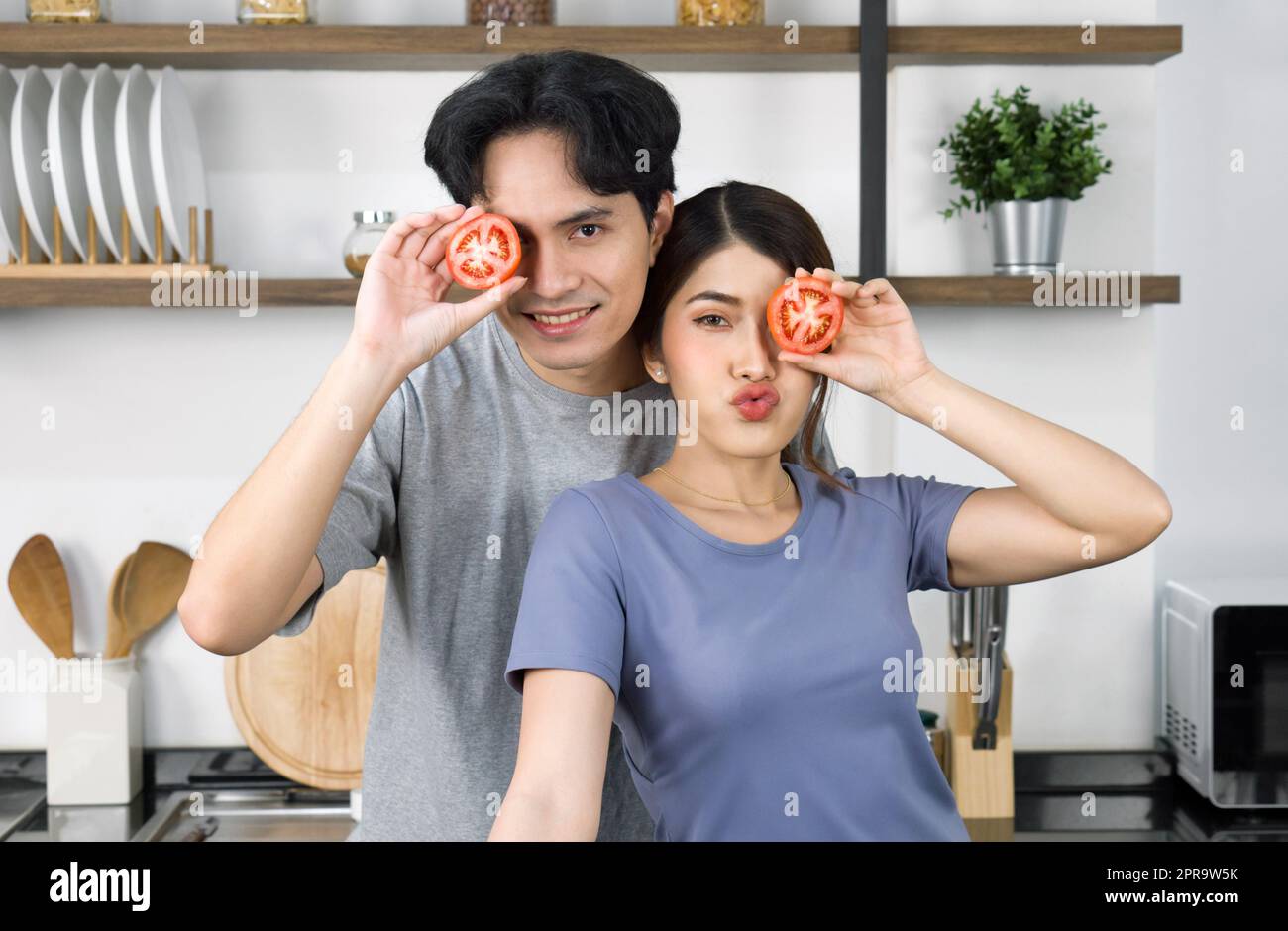 Young couple in casual clothe, hold slice tomato in eye position. Asian lover spend morning time together in the kitchen. Stock Photo