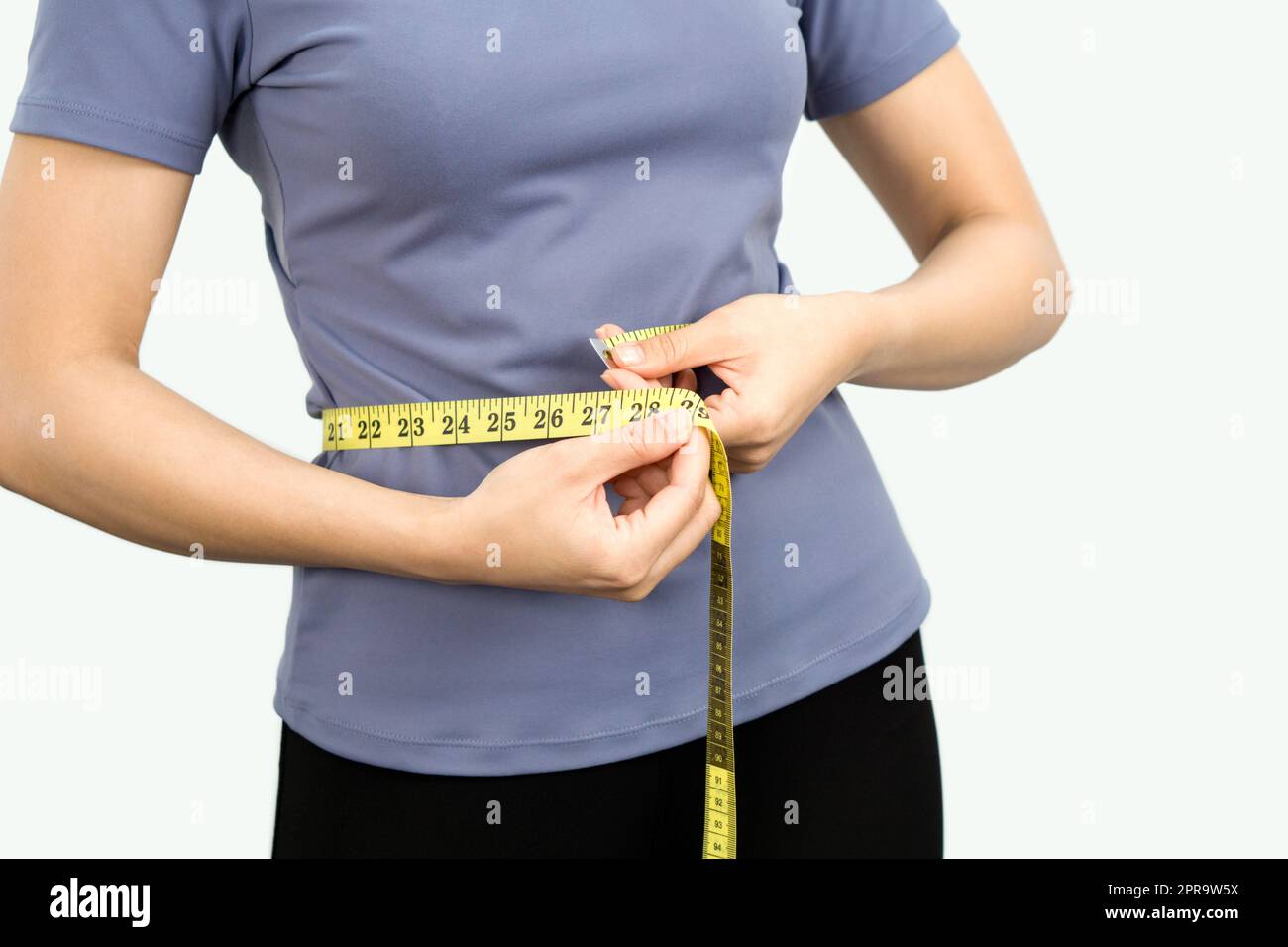 Closeup female hands in exercise clothes use a tape measure to measure around the waist. Healthy nutrition and weight losing concept. Portrait on white background with studio light. Stock Photo