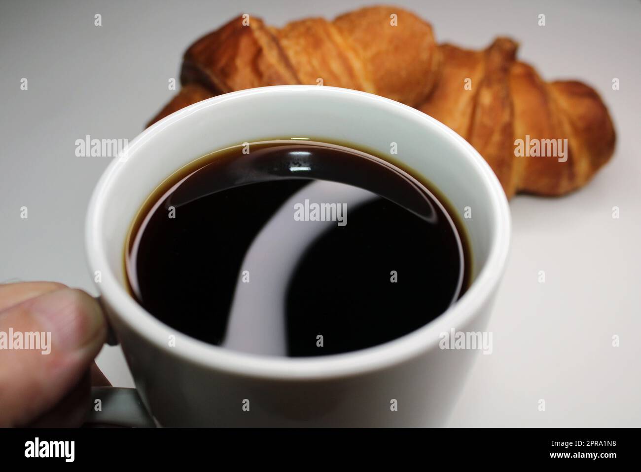 White cup with black coffee close-up on a light background. There are two croissants in the background. Cooking, home baking Stock Photo