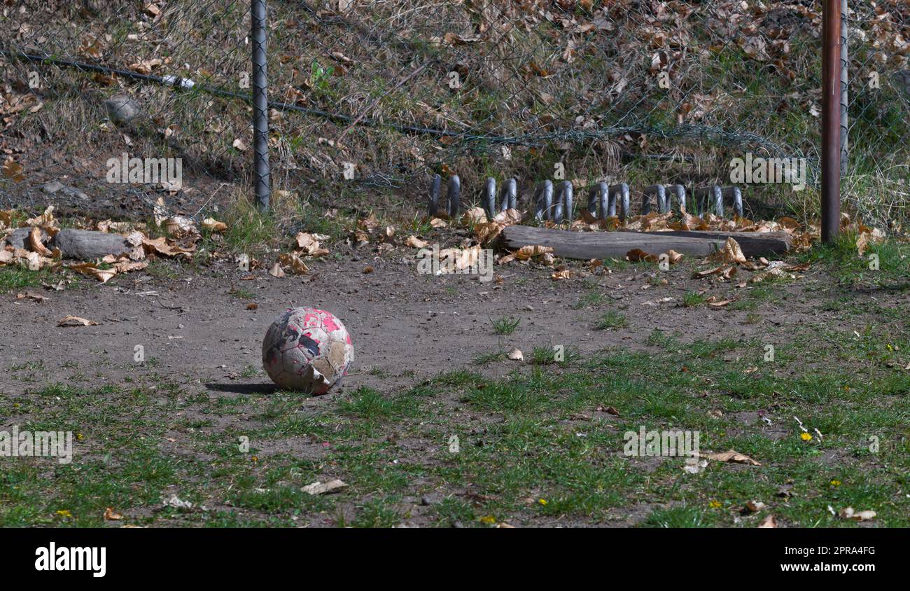 Old dirty football lying on the abandoned playground. Parts of the old fence, post and goal are visible and show the abandoned state of the place. Stock Photo