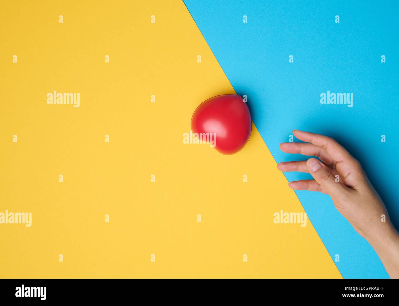 Red heart and a woman's hand reaches for it on a blue-yellow background Stock Photo