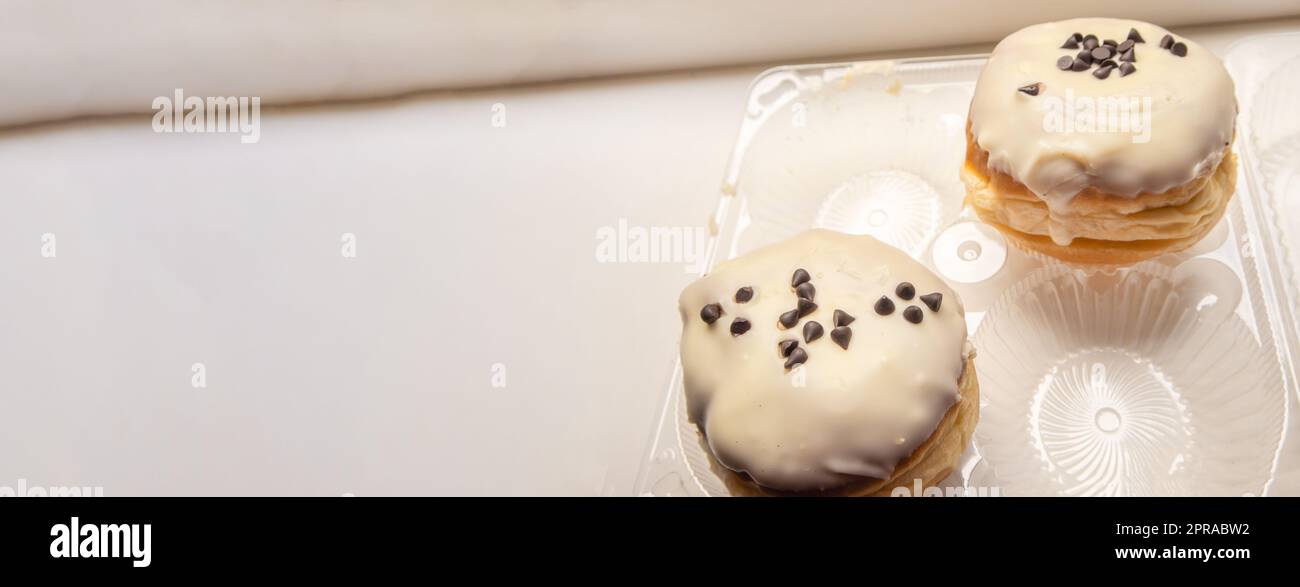 Two delicious donuts with icing sugar and chocolate chips on a white background in a plastic transparent container, a banner with a copy of the space on the left Stock Photo