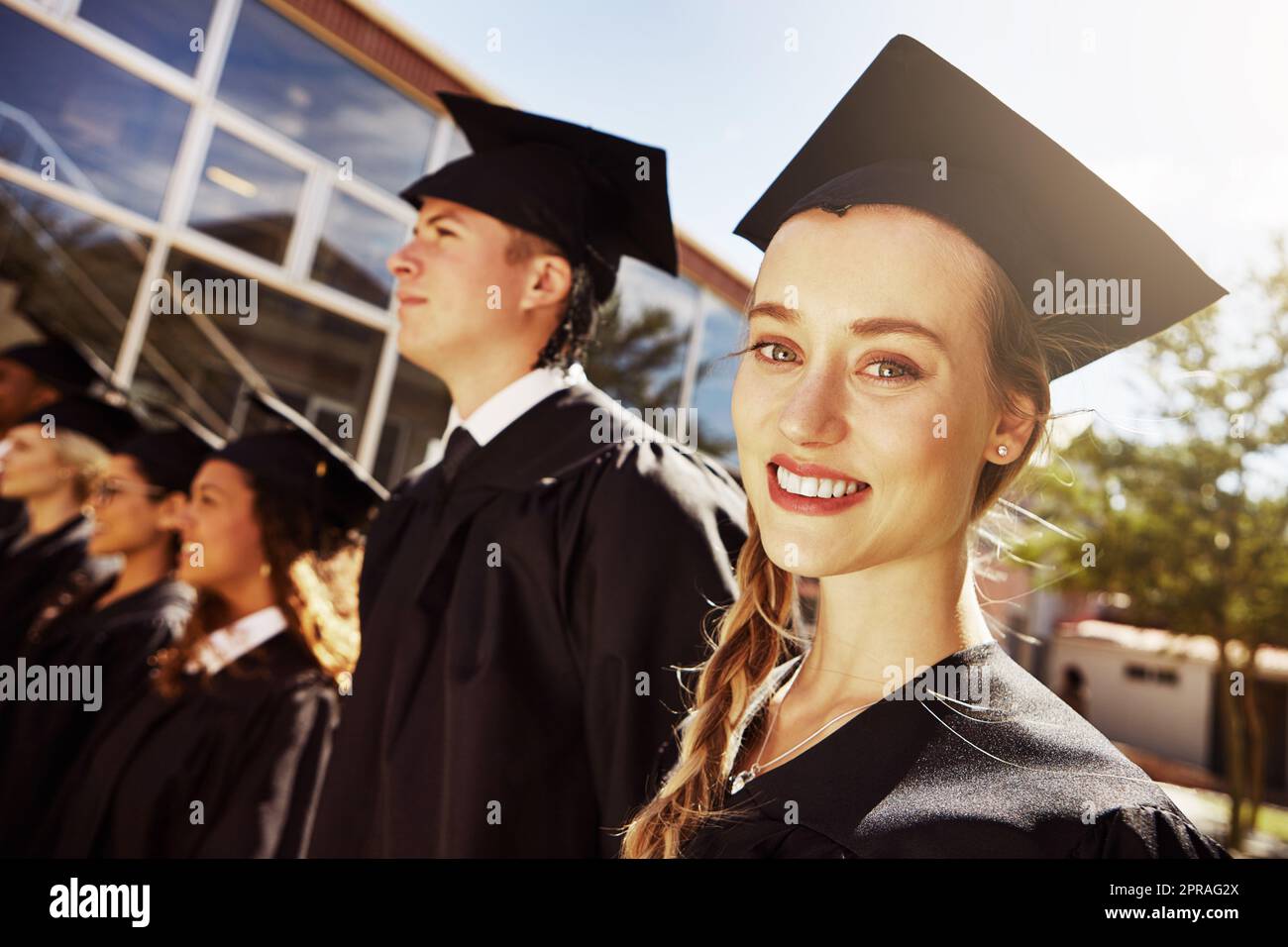 She has a bright future ahead of her. Portrait of a smiling university student on graduation day with classmates in the background. Stock Photo