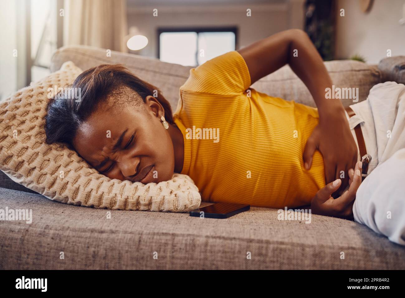 Sad and sick girl with stomach pain, period cramps or menstruation rubbing her belly ache on sofa. One unhappy woman suffering from endometriosis discomfort in abdomen due to PMS and bad healthcare Stock Photo