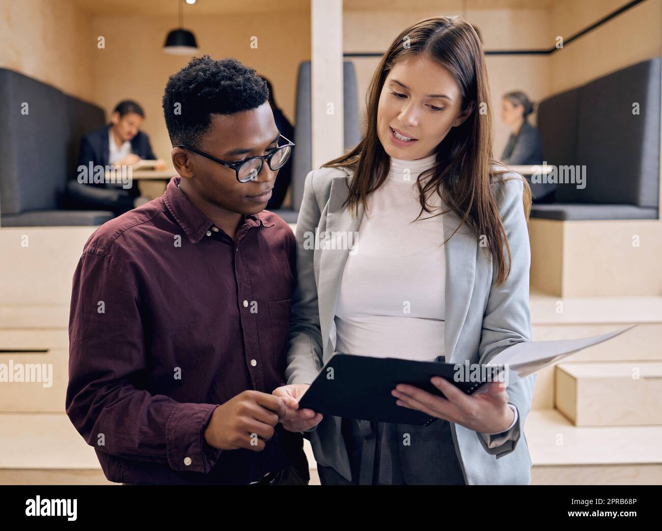 Its great to have a colleague you can rely on. two young businesspeople looking over paperwork with their colleagues in the background. Stock Photo