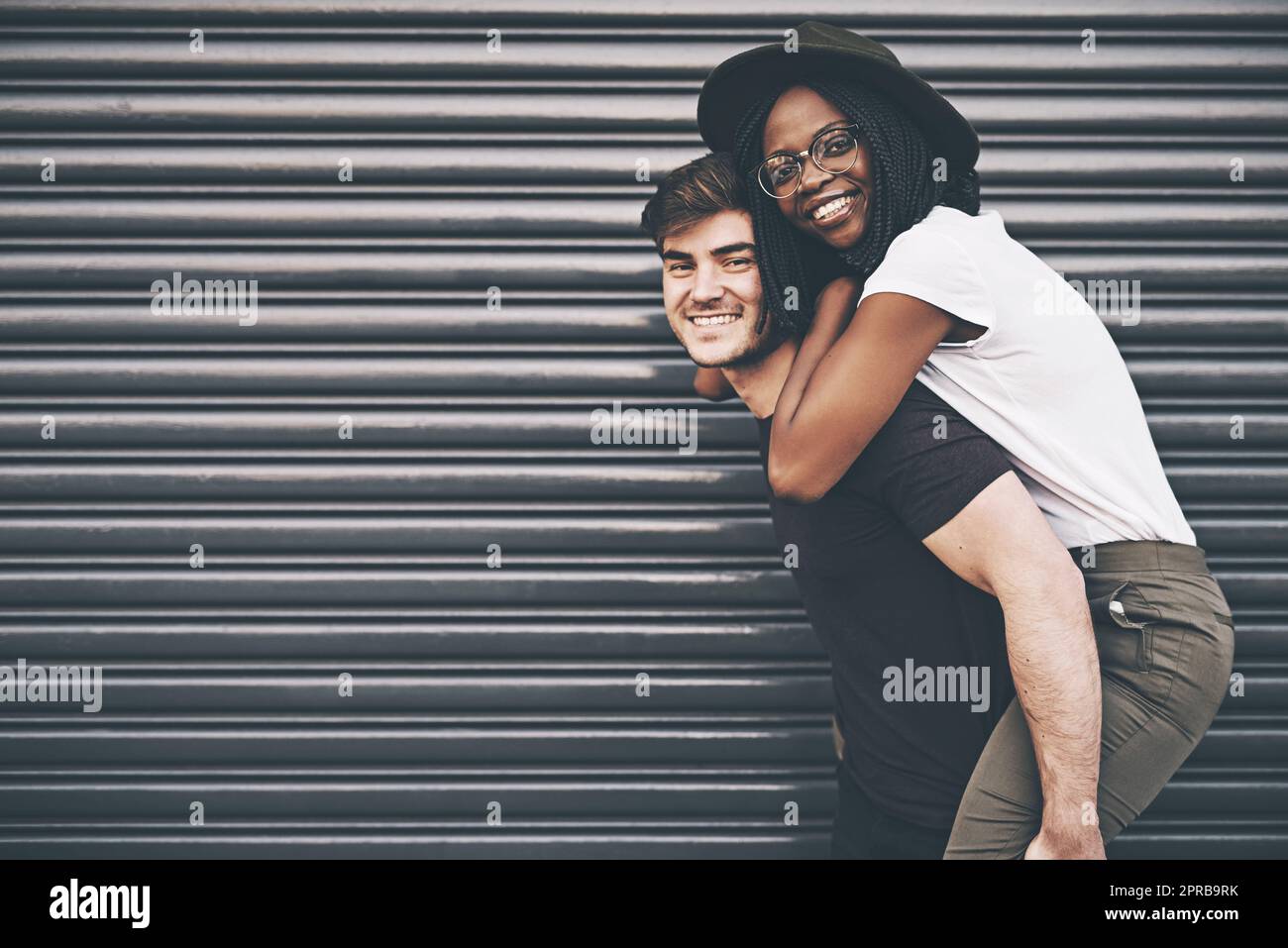 Cute, loving and affectionate interracial couple having fun, being playful and enjoying time together on copy space background. Happy boyfriend giving young girlfriend a piggyback ride outside Stock Photo