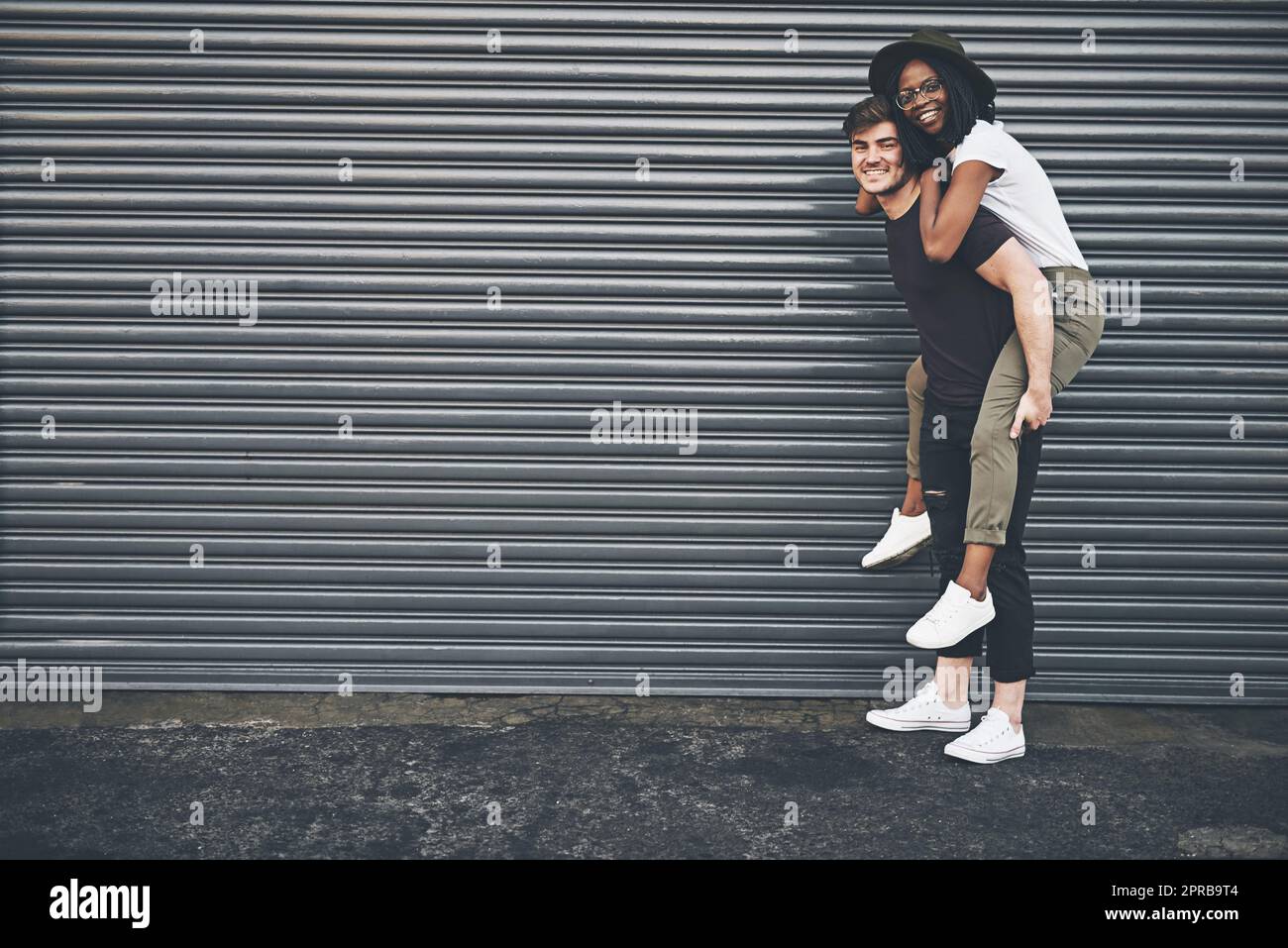 Fun, trendy and happy interracial couple enjoying a piggyback ride together outside against an urban background. Lovers bonding and being playful. Smiling and relaxing while walking outdoors Stock Photo