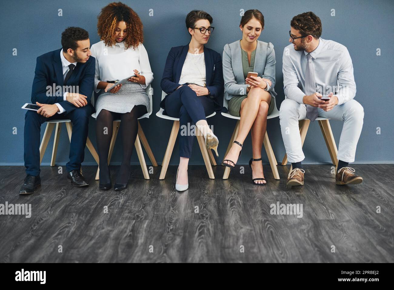 Group of business people networking on phones, digital tablets and social media, waiting in line. Diverse team of colleagues sitting in queue, talking and discussing against blue copyspace wall. Stock Photo