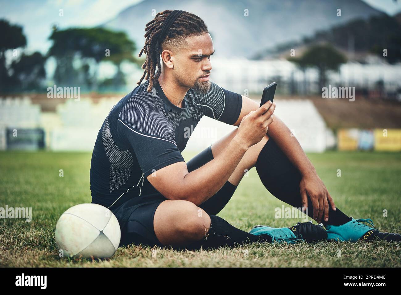 Im pretty sure my teammates should be here by now... Full length shot of a handsome young rugby player using a smartphone while sitting in the playing field during the day. Stock Photo