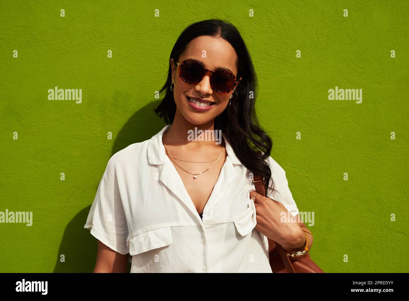 Living life the way I want to. Cropped portrait of an attractive young woman wearing sunglasses while standing alone against a green background in the city. Stock Photo