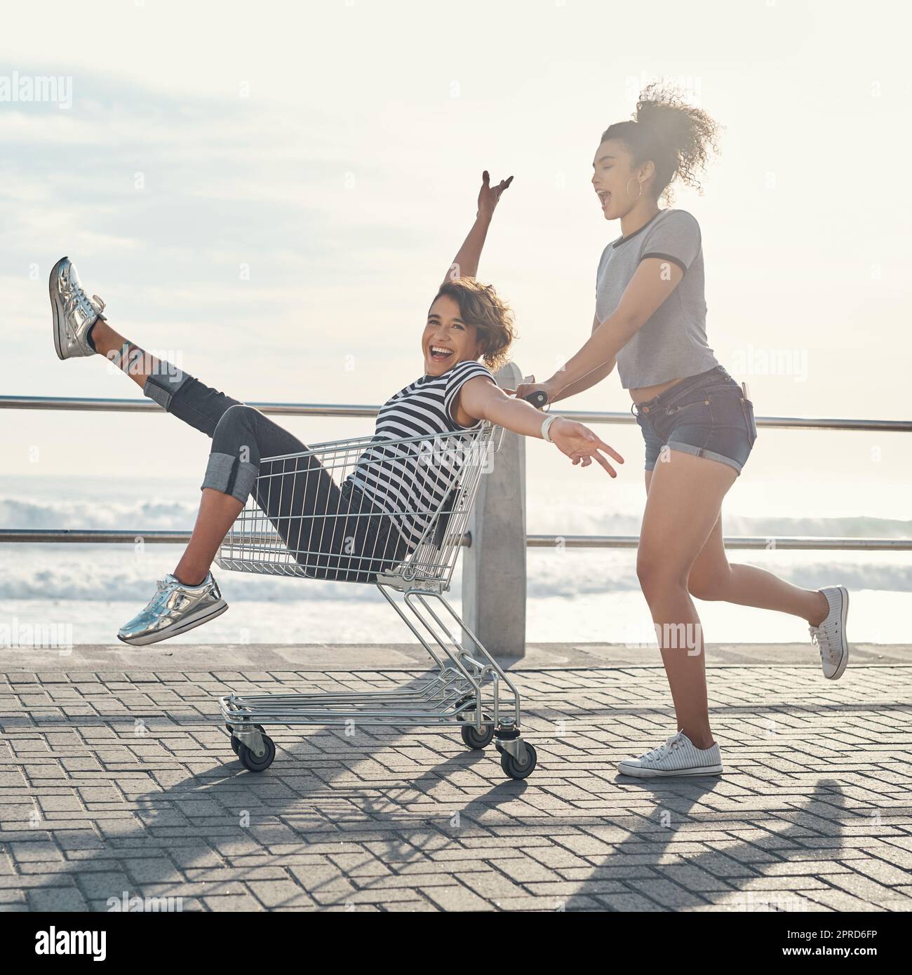 Good friends is the greatest gift. a young woman pushing her friend around on the promenade in a shopping chart. Stock Photo