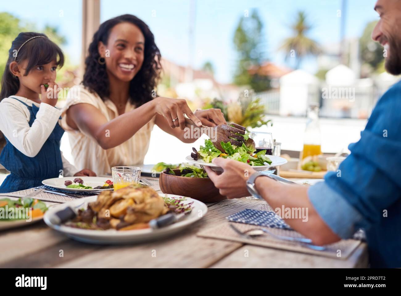 She always makes sure we maintain a healthy diet. a beautiful young woman dishing up salad on her husbands plate while enjoying a meal with family outdoors. Stock Photo