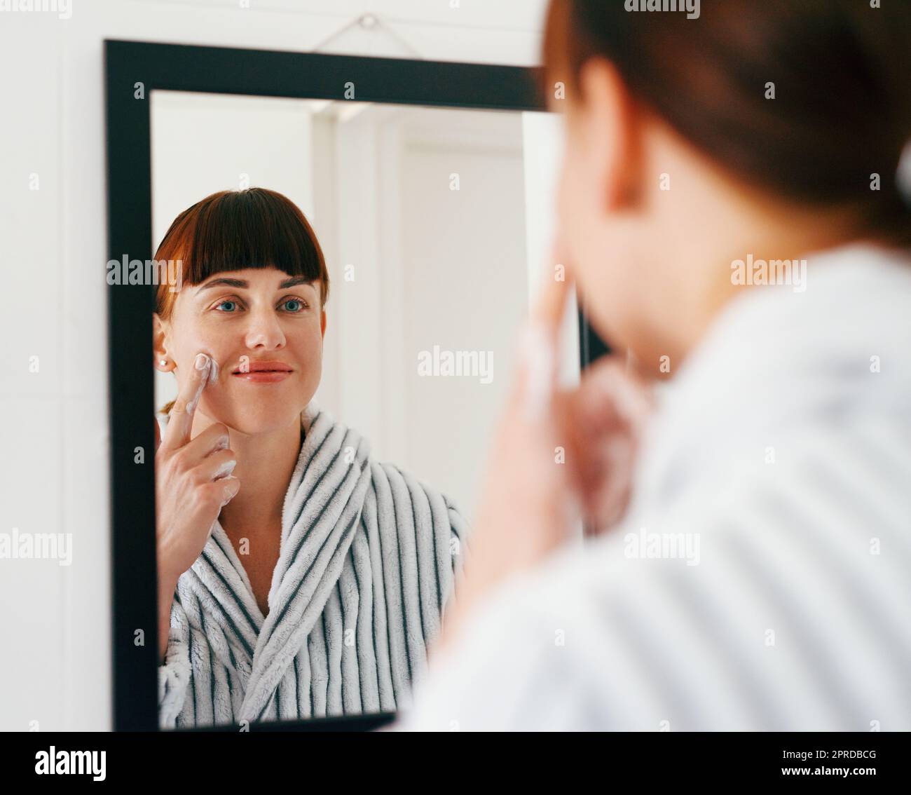 Your beautiful face deserves all the pampering it can get. an attractive young woman applying moisturizer on her face while standing in front of the mirror at home. Stock Photo