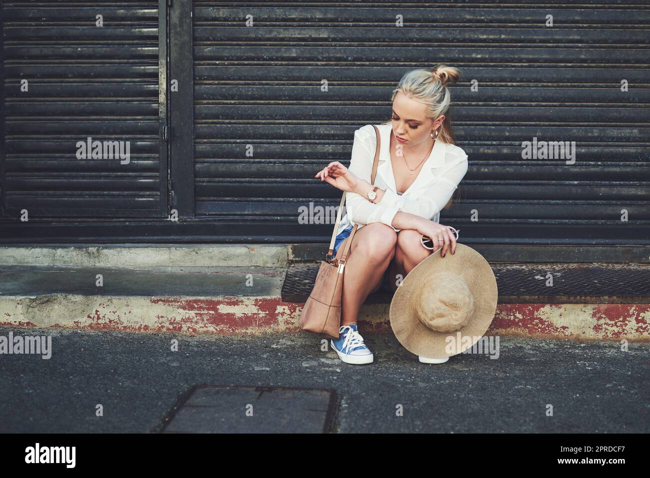 I cant believe Im still waiting here. Full length shot of an upset young woman checking the time on her wristwatch while sitting outdoors. Stock Photo