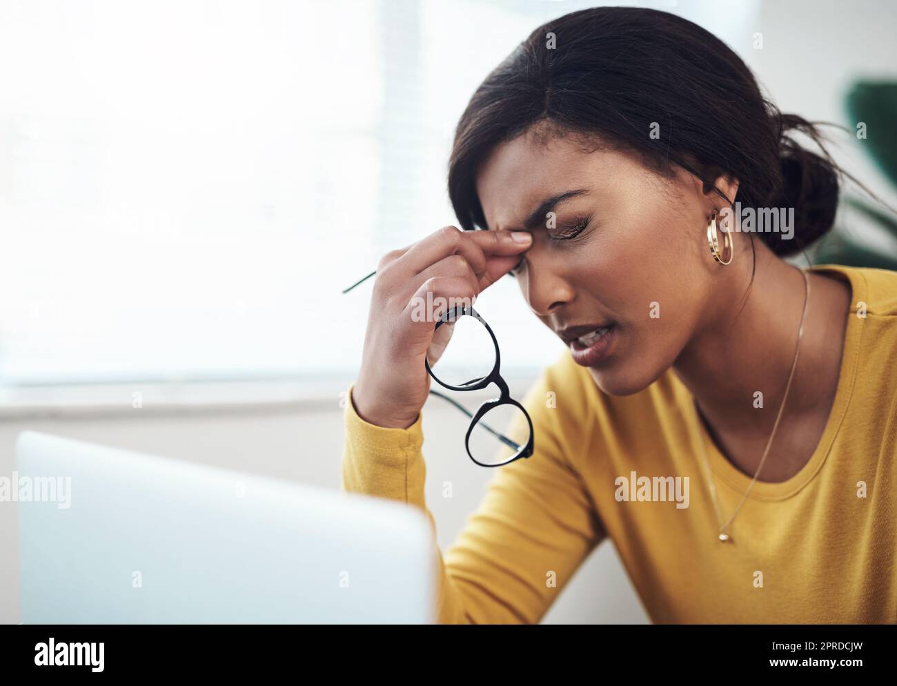 This headache came out of nowhere. an attractive young woman sitting alone at home and suffering from a headache while using her laptop. Stock Photo
