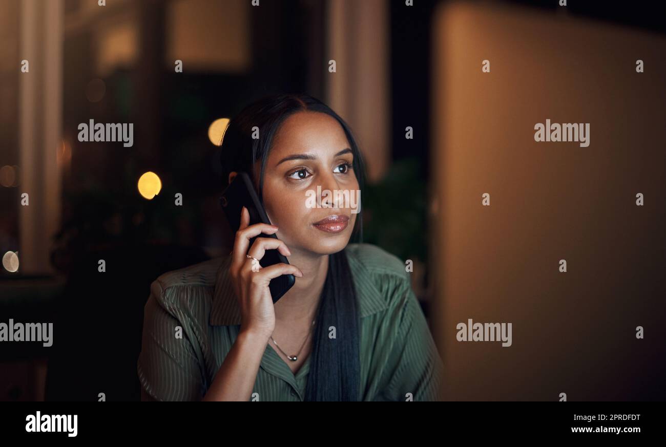 Sometimes you just have to rise early and work late. a young businesswoman talking on a cellphone while using a computer in an office at night. Stock Photo