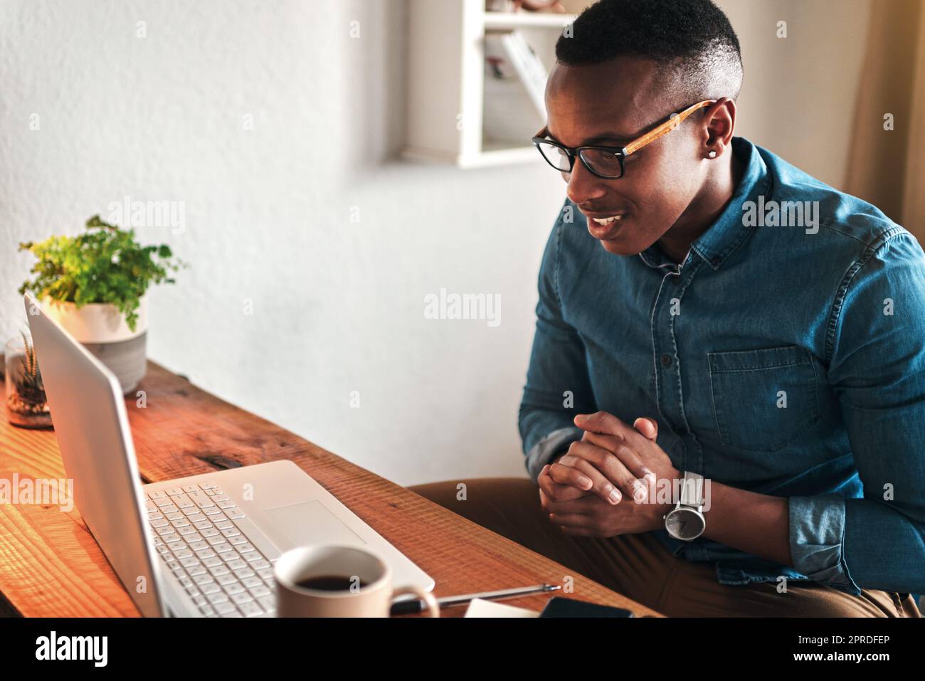 Male online worker on his computer working freelance sitting in a home office alone. Casual digital employee reading and looking at tech and web research. Young modern man doing a remote tech job Stock Photo