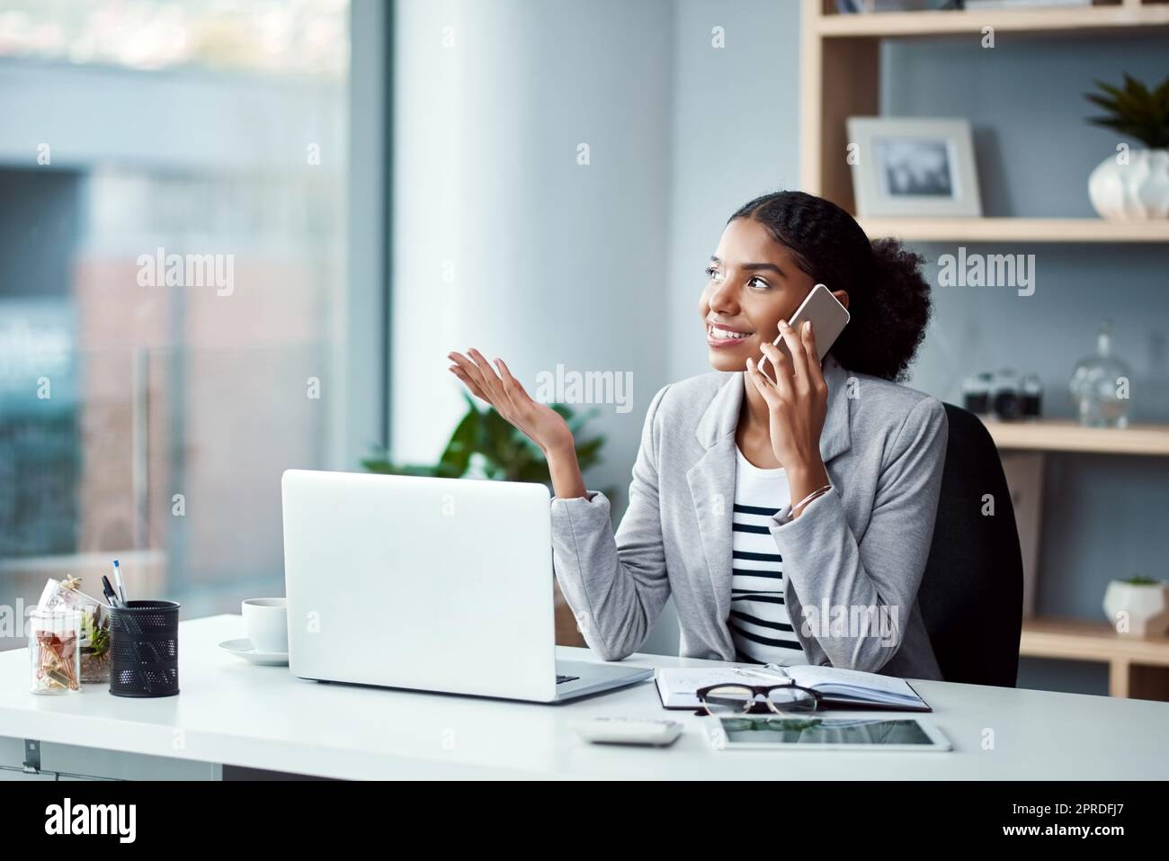 Young business woman on a phone call talking and planning while sitting alone at a desk on a office computer. Female worker discussing work on her smartphone. An employee embracing modern technology Stock Photo