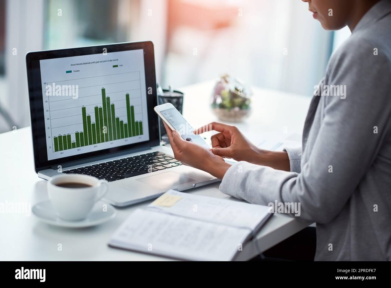 Female data analyst or finance manager working on phone in a modern office. Business professional multitasking with graph and chart data on laptop, planning and analysis at the workplace. Stock Photo