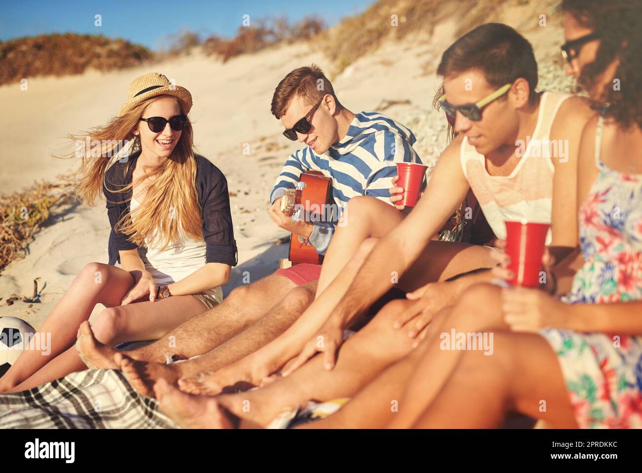 Hes the kind of friend thatll play you a song. a handsome young man playing a guitar for his friends on a summers day at the beach. Stock Photo