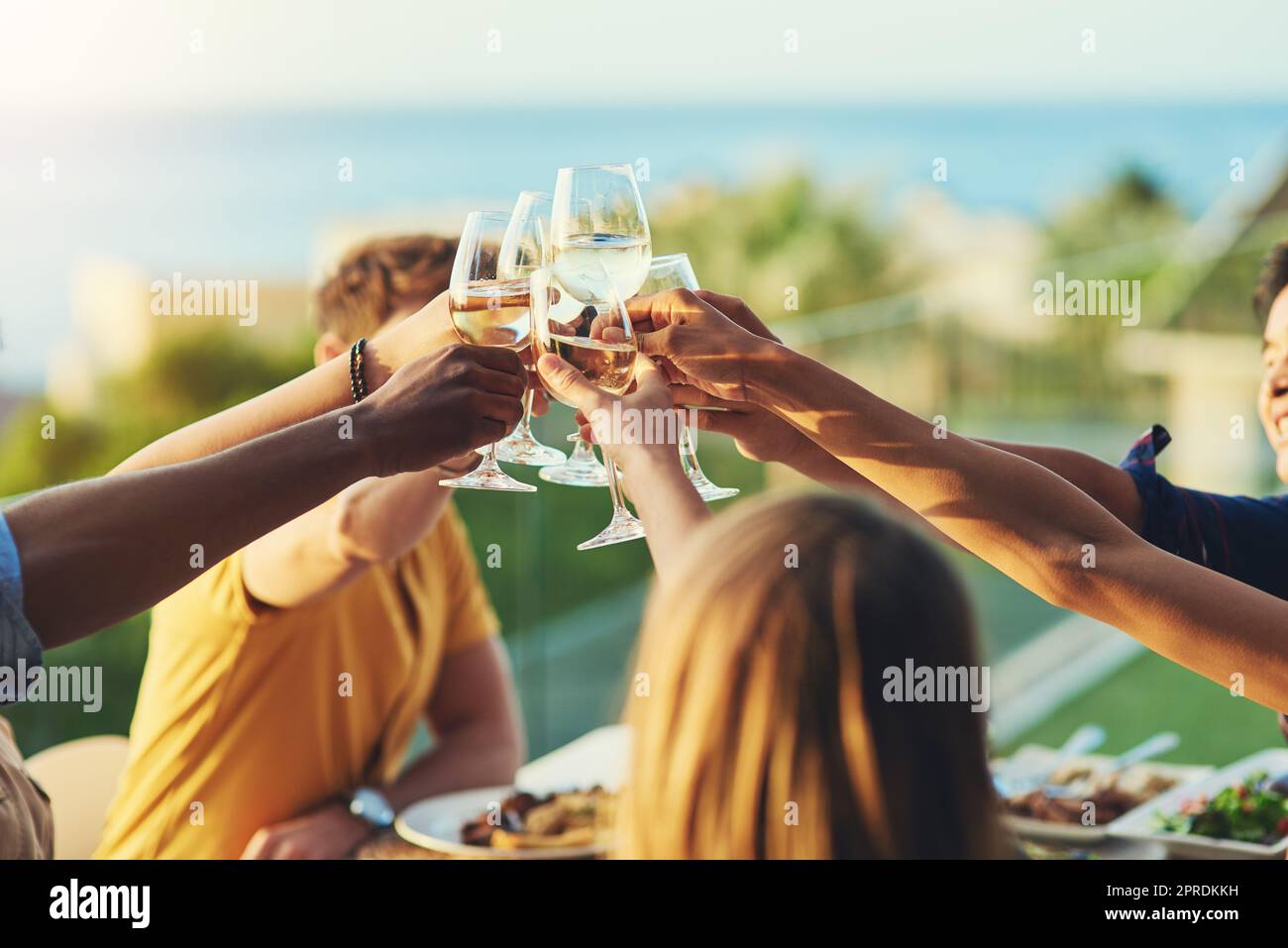 To us growing closer and stronger together. a group of friends raising up their glasses for a toast while sitting around a table together outdoors. Stock Photo