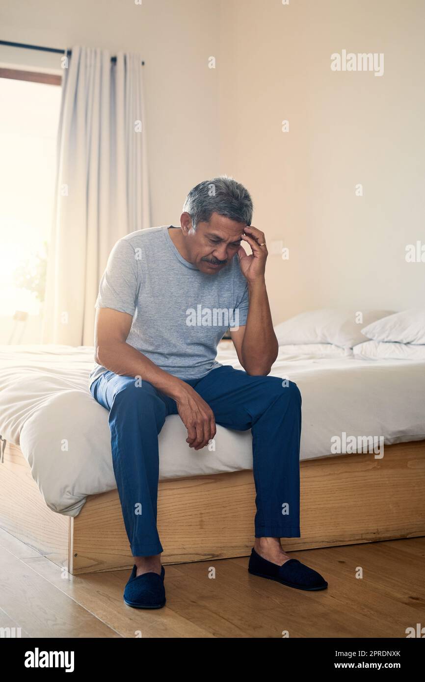 This headache is getting really irritating. a stressed out mature man holding his head in discomfort while being seated on his bed at home. Stock Photo