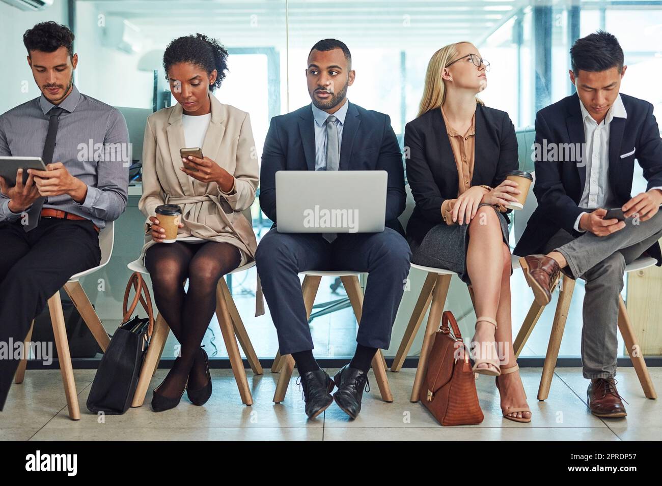 a group of focussed young businesspeople seated together on chairs and making notes using different methods while waiting in the office during the day Stock Photo