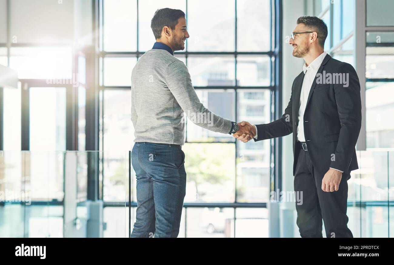 Businessman standing, smiling and with a handshake, greets a colleague in an office. Executive meets client or partner and shakes hands. Two happy professionals agree on a successful business project Stock Photo