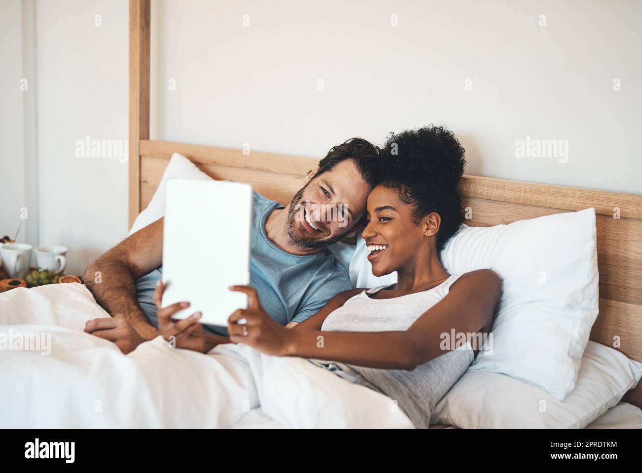Happy couple taking selfie on a tablet in bed together after waking up. Interracial husband and wife enjoying their relationship, having fun while bonding and relaxing indoors on a weekend Stock Photo