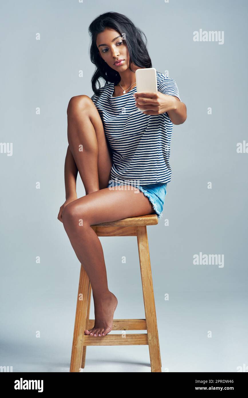 Her angles are on point. Studio shot of a beautiful young woman taking a selfie while sitting on a wooden stool. Stock Photo