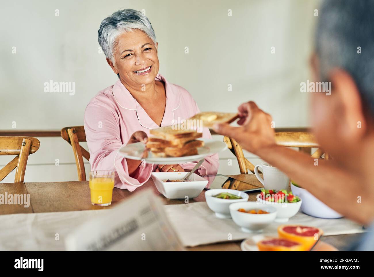 Would you like another one. a cheerful elderly woman handing over a plate of toast to her husband during breakfast at home. Stock Photo
