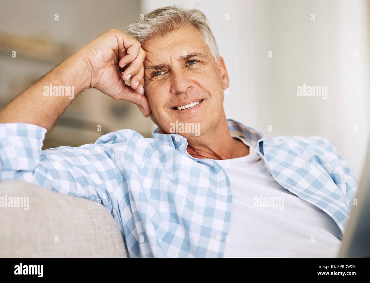 Handsome mature man relaxing on a sofa, casual, carefree and happy at home. Senior male enjoying the pleasure of retired life, satisfied and stressless. Older guy thinking, smiling and daydreaming Stock Photo
